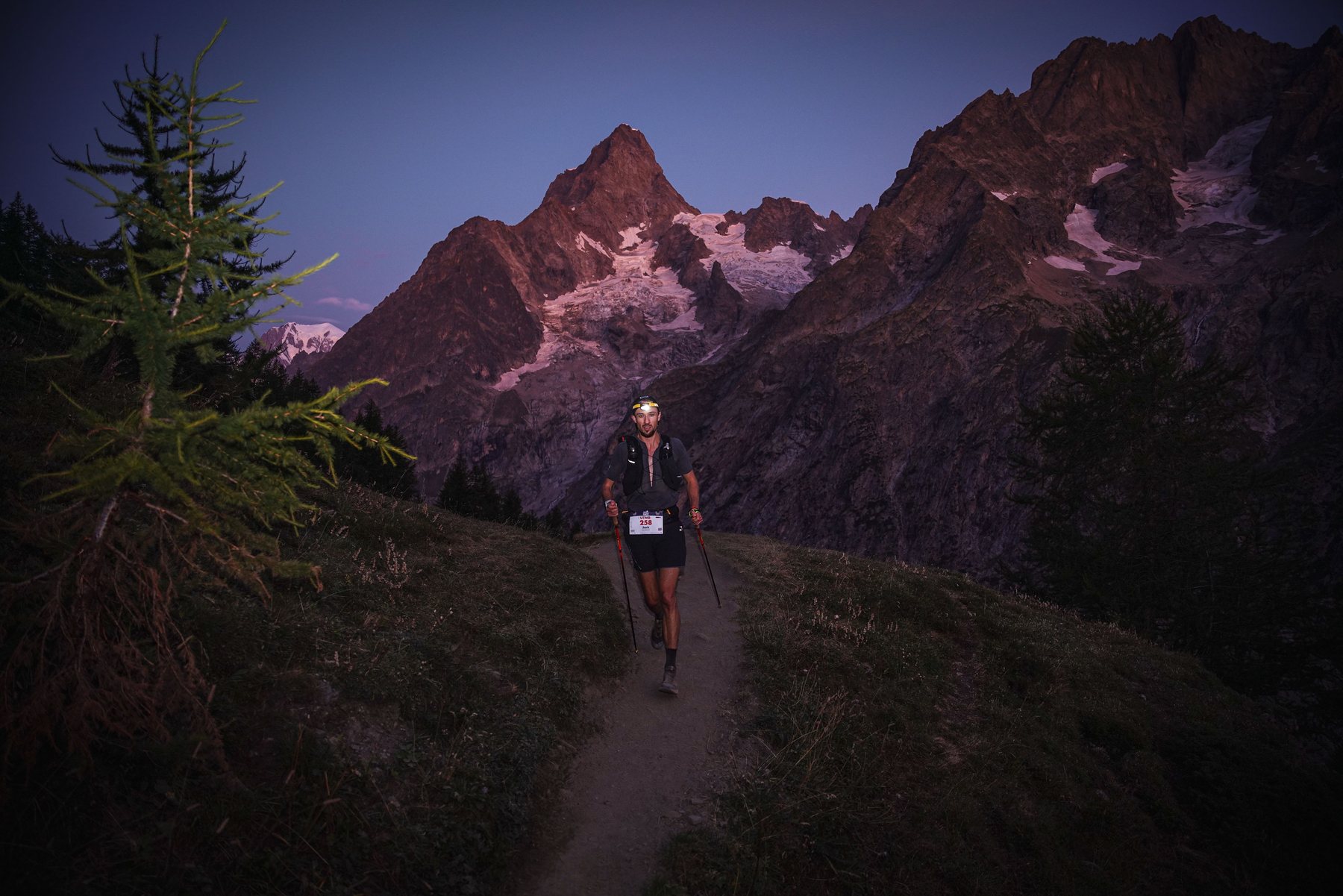 Jack Chamberlain steigt bei der UTMB Mont-Blanc im ersten Licht mit Stirnlampe einen Pfad hinauf, dahinter die rosa beleuchtete Aiguille Noire.